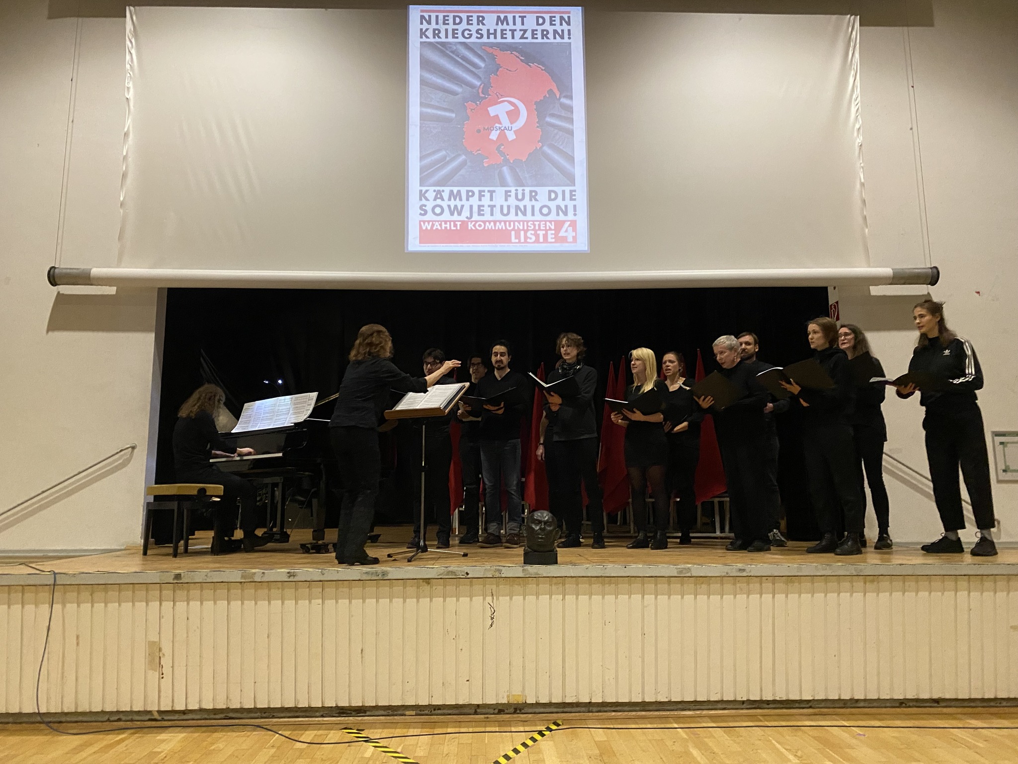 Foto vom Chor auf der Bühne beim Singen. Der Chor ist in schwarz gekleidet, im Hintergrund sind rote Fahnen und die Projektion eines kommunistischen Wahlplakats zu sehen.