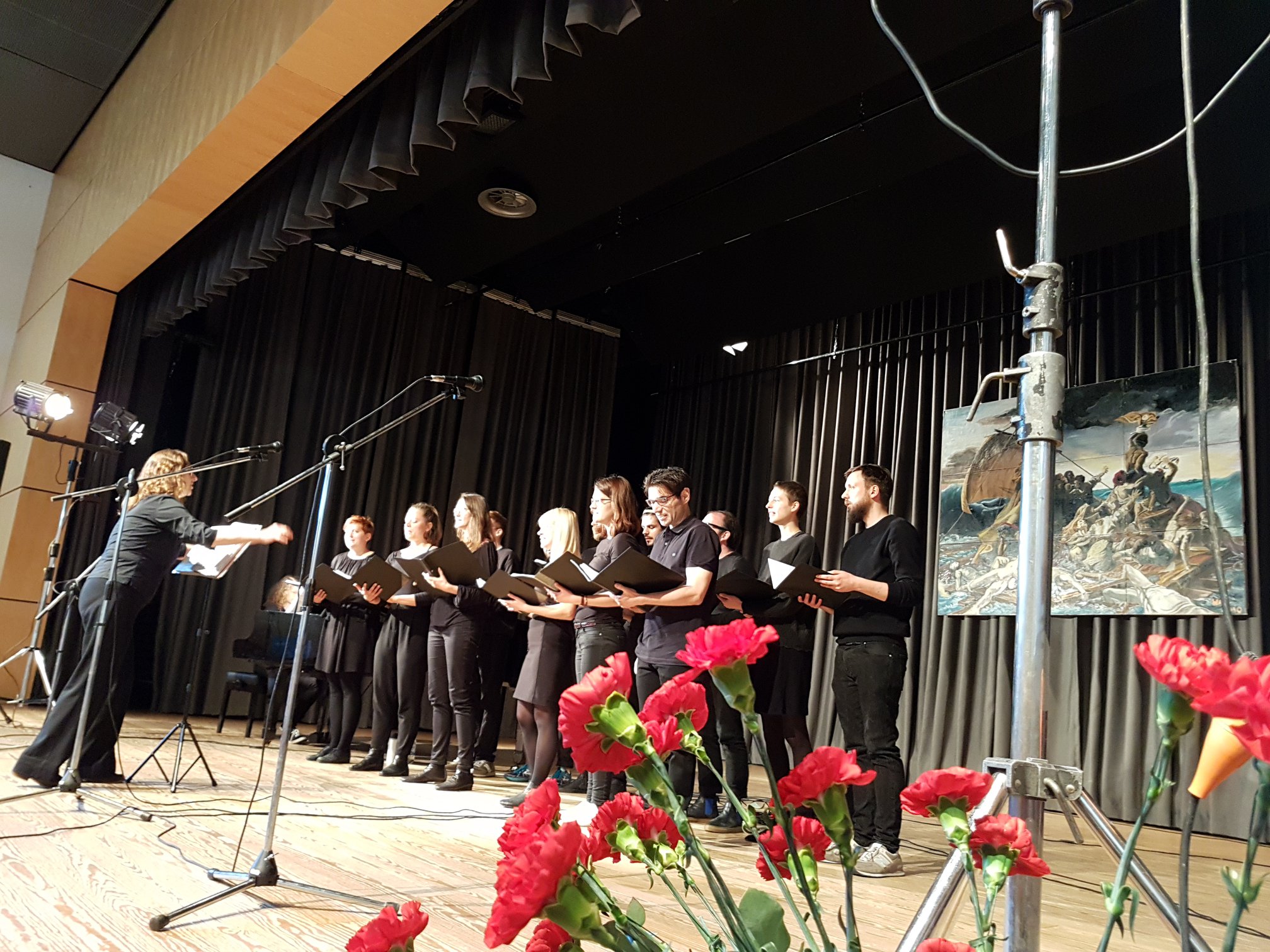 Der Chor ist in schwarz gekleidet auf einer großen Bühne beim Singen zu sehen. Im Vordergrund sind rote Nelken zu sehen.