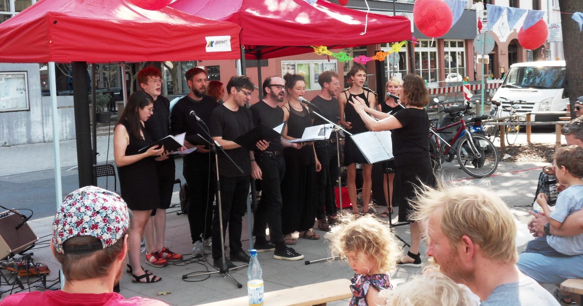 Foto vom Chor beim Singen auf der Straße unter roten Pavillons. Im Vordergrund ist Publikum zu sehen.