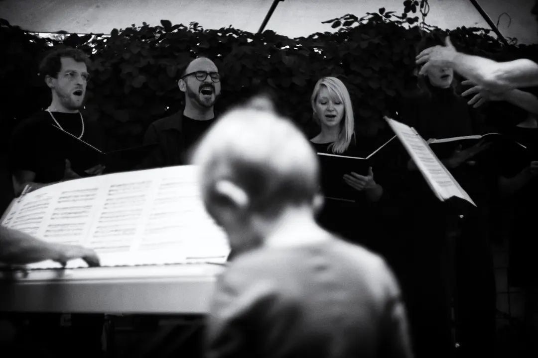 Schwarzweiß-Foto von Mitgliedern des Chors beim Singen. Am linken Bildrand ist ein Klavier zu sehen, am rechten Bildrand ragen dirigierende Hände hinein.